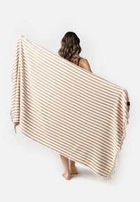 Gestreepte deken in beige en crème, met een zachte textuur, franjes langs één rand en een rechthoekige vorm.