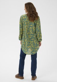 Långärmad tunika i grön med mönster av gula löv, lättviktig tyg, rund nederkant, kombinerad med mörkblå jeans och bruna skor.
