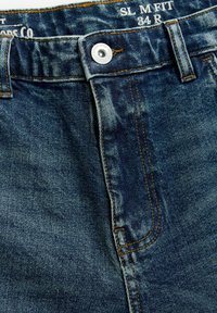 Jeans din denim albastru cu o textură ușor decolorată, prezentând cusături galbene, un sistem de închidere cu buton argintiu și un etichetă care indică mărimea 34 R.