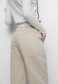 Beige bomullbyxor med elastisk midja, med överdimensionerade bakfickor och en avslappnad passform. Tyget har en slät yta.