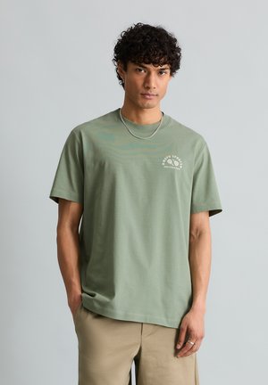 Jonge man met krullend haar draagt een lichtgroen T-shirt met "Mood Sports" opdruk en beige broek, staat met één hand in zijn zak tegen een grijze achtergrond.