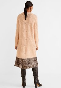 Vaaleanpunainen neulottu neuletakki, jossa on kaapelikuvio, kerroksittain leopardikuosisen hameen päällä, yhdistettynä polven korkuisiin mustiin saappaisiin.