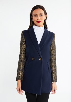 Donna con capelli lunghi e scuri indossa un cappotto doppiopetto blu navy con dolcevita bianco e maniche lunghe con motivo dorato, in piedi davanti a uno sfondo bianco.
