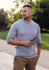 Gray long-sleeve v-neck sweater, tan pants, and holding a gray wallet with a zipper, featuring a black card slot. Natural outdoor setting.