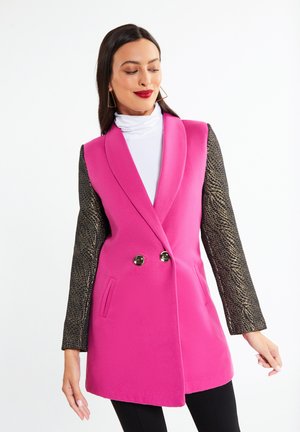 Femeie cu blazer roz aprins fără mâneci cu mâneci lungi cu model auriu, helancă albă, pantaloni negri și cercei geometrici pe fundal alb.