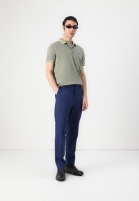 Polo shirt de culoare verde măsliniu, asortat cu pantaloni bleumarin. Modelul poartă ochelari de soare negri și bocanci negri până la gleznă. Design simplu și ajustat.