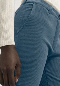 Blå kordbyxor med vertikal ribbad textur, utrustade med sidofickor och knappstängning, kombinerat med en ljusfärgad ribbad tröja.