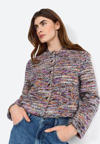 Texturierte, mehrfarbige Jacke in Lila-, Orange- und Grautönen, mit Kragen, Knöpfen und langen Ärmeln, kombiniert mit Jeans.