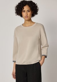 Frau mit lockigem dunklem Haar, die ein beiges Langarmshirt mit schwarzen gestreiften Bündchen und dunkel gestreifte Hose trägt, steht vor einem schlichten Hintergrund.