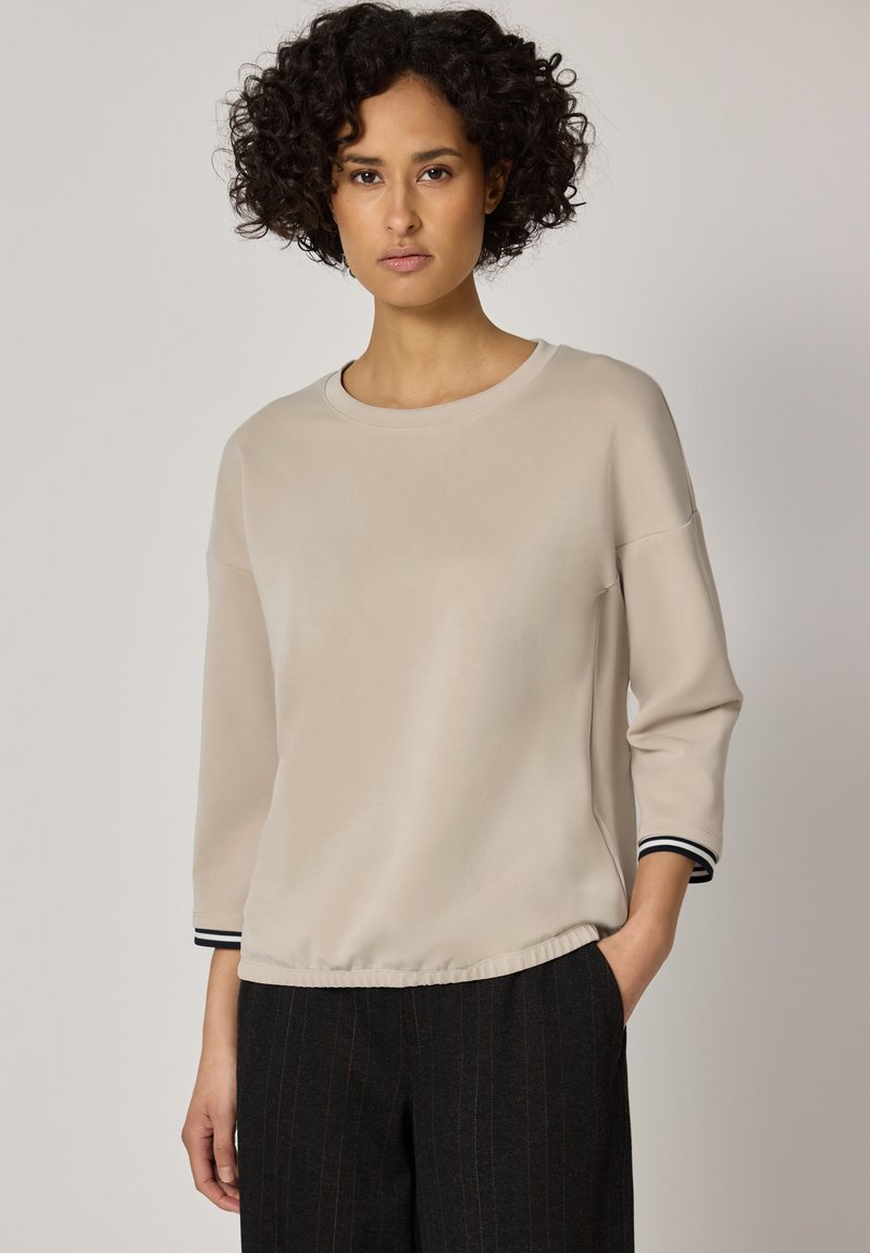 Frau mit lockigem dunklem Haar, die ein beiges Langarmshirt mit schwarzen gestreiften Bündchen und dunkel gestreifte Hose trägt, steht vor einem schlichten Hintergrund.