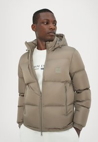 Puffer jacket in light brown, featuring a hood, zippered front, and elastic cuffs. Textured, quilted design with logo detail on chest.