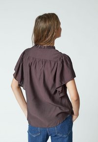 Blusa marrone a maniche corte con collo a volant e vestibilità ampia, caratterizzata da un dettaglio plissettato sulla parte posteriore. Abbinata a jeans in denim blu.