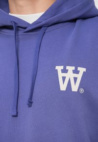 Fialová mikina s kapucňou z bavlny; na prednej strane má biele logo s stilizovaným „W“, šnúrky pri krku a textúrovanú tkaninu.