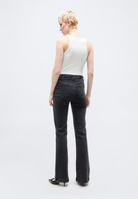 Ξανθό άτομο που στέκεται πλάγια, φοράει λευκή αμάνικη μπλούζα, ξεβαμμένο μαύρο τζιν με ψηλή μέση και μαύρα παπούτσια με μικρό τακούνι.