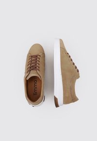 Zapatillas de ante beige con cordones marrones, suelas de goma blanca y una textura suave. Logo visible en las plantillas. Diseño clásico de bajo perfil.