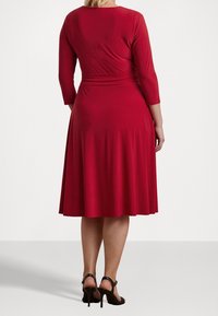 Vestido rojo de longitud a la rodilla con un cuerpo entallado y falda acampanada. Mangas largas y tela suave y elástica. Llevado con sandalias negras de tacón alto.