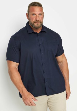 Muskelmand med skæg iført mørkeblå kortærmet skjorte med knapper og beige bukser, stående mod en hvid baggrund.