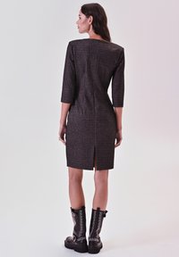 Eng anliegendes braunes Kleid mit strukturiertem Muster, dreiviertel Ärmeln und einem Rückenschlitz, kombiniert mit schwarzen, mit Nieten besetzten Knöchelstiefeln.