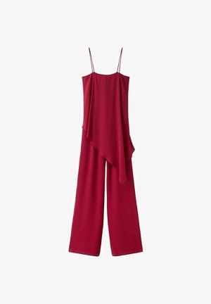 Žena s tmavými vlasmi v voľnom bordovom bezrukávovom overale so širokými nohavicami stojaca pred zodpovedajúcim ružovým pozadím so založenými rukami.