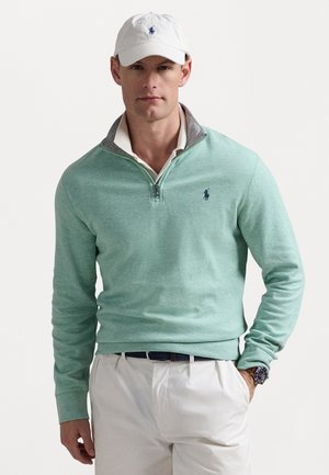 Pull léger vert clair à fermeture éclair quart, avec un col gris et un logo bleu, associé à une chemise blanche, une casquette blanche et un pantalon de couleur claire.