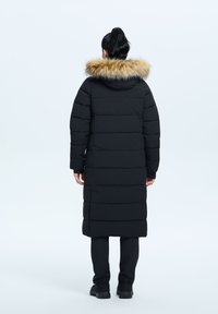Schwarze Pufferjacke mit langem Design und Kapuze mit Kunstfellbesatz. Sie hat horizontale Steppung und lange Ärmel und wird mit schwarzen Hosen und Schuhen kombiniert.