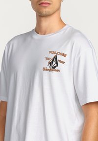 T-shirt en coton blanc à manches courtes avec un design graphique orange et noir, portant le texte "VOLCOM Workwear" sur la poitrine gauche.