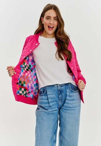 Prešívaná ružová bunda s farebnou károvanou podšívkou, nosená cez bielu košeľu a svetlomodré džínsy.