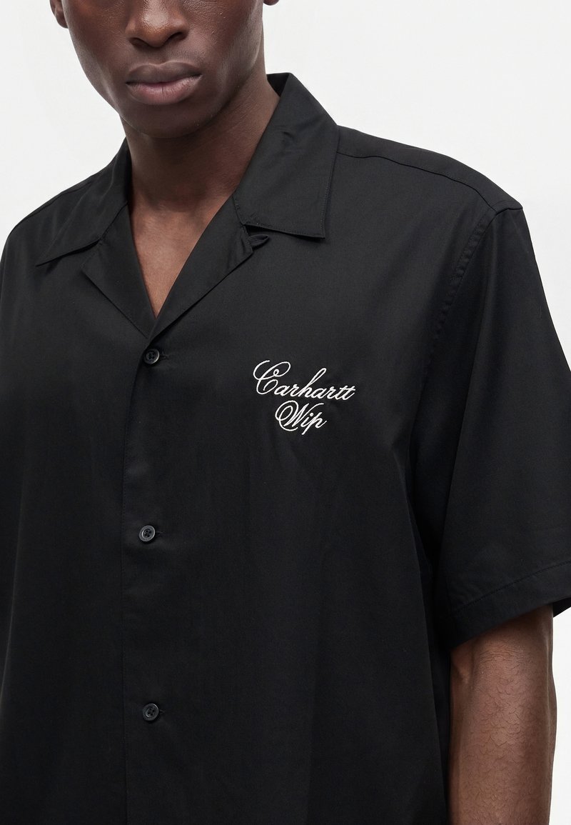 Uomo che indossa una camicia nera a maniche corte con bottoni e scritta bianca "Carhartt Wip" sul petto.