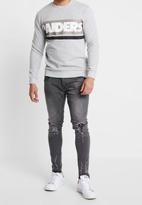 Sweat-shirt gris à col rond avec un graphique "RAIDERS", associé à un jean skinny gris foncé usé et des baskets blanches.