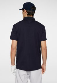 Marinblå polo-shirt med en strukturerad krage och korta ärmar. Baksidan har en logotyp vid nacklinjen. Matchas med ljusa byxor och en keps.