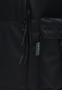 Schwarzer Rucksack aus Stoff mit strukturierter Oberfläche, Reißverschluss, Kordelzug-Akzenten und kleinem grünen Lacoste-Logo-Tag.