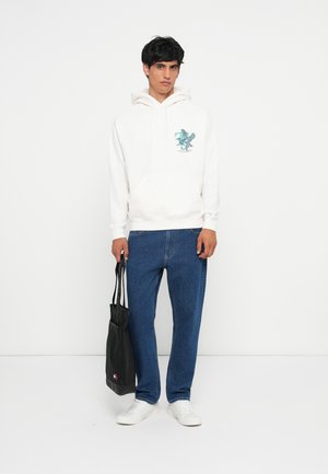 Witte hoodie met een teal graphic, gecombineerd met blauwe jeans en witte sneakers. Het model houdt een zwarte totebag vast.