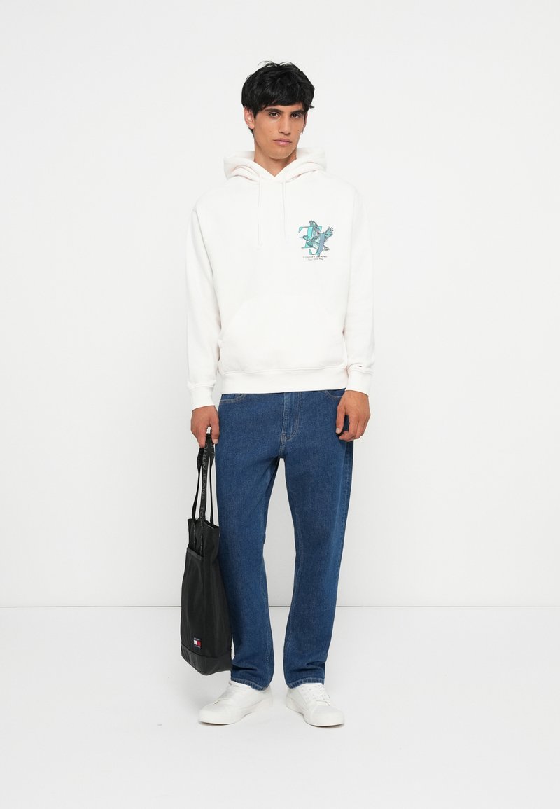 Hvit hettegenser med en teal grafikk, kombinert med blå jeans og hvite joggesko. Modellen holder en svart tote bag.
