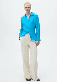 Camicia di lino turchese con vestibilità rilassata, caratterizzata da colletto e chiusura a bottoni. Abbinata a pantaloni a gamba larga crema e sandali neri.
