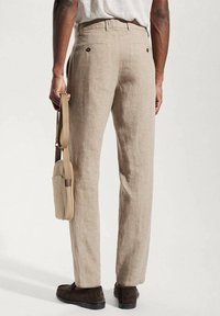 Světle béžové lněné kalhoty se střihovým designem rovného nohavičkového, s dvěma zadními kapsami, nošeny s šedou košilí a drženy hnědou křížovou taškou.