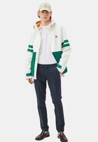 Giacca con cappuccio bianca e verde con fodera arancione, abbinata a una camicia bianca e pantaloni blu scuro. Scarpe marroni lucide completano l'outfit.
