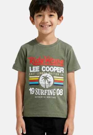 Jeune garçon souriant portant un t-shirt vert olive à thème surf "Ride Wave Lee Cooper" avec un palmier et des rayures colorées, debout.
