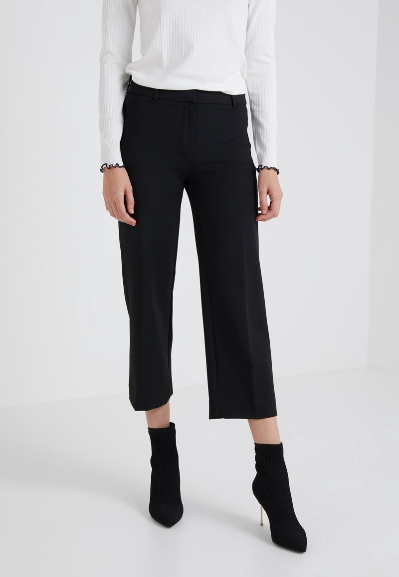 Femme portant un pantalon large noir court, des bottines noires à talons et un haut blanc à manches longues avec des bords à volants aux poignets.