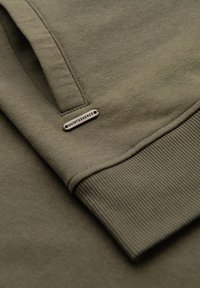 Dettaglio di una felpa verde oliva, realizzata in morbido cotone, con polsini a coste e una piccola etichetta metallica con la scritta "QUINTESSENCE."