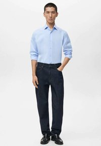 Light blue button-up shirt with long sleeves paired with dark denim jeans. Black shoes. Neutral background. Simple, classic design.