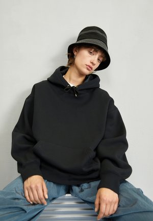 Sudadera negra de gran tamaño con bolsillo canguro, detalle de cordón y puños acanalados, combinada con un sombrero tipo cubo negro con acentos blancos.