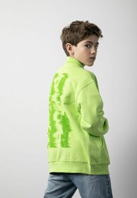 Neonowy zielony sweter z wysokim kołnierzem i teksturowanymi akcentami na plecach, w zestawie z jasnoniebieskimi dżinsami. Gładki materiał, luźny krój.