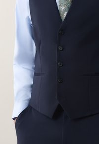 Man wearing navy blue buttoned vest over light blue shirt with floral patterned green tie, hand in pocket.