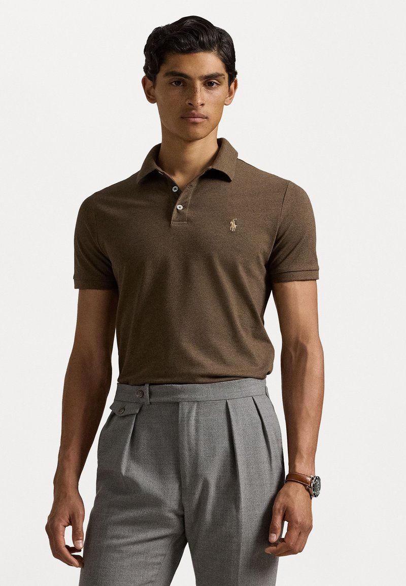 Polo shirt marron à manches courtes avec col et petit logo ; porté avec un pantalon plissé gris clair et une montre à bracelet en cuir.