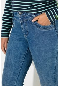 Lähikuva henkilöstä, joka pukeutunut sinisiin farkkuihin ja raidalliseen pitkähihainen paitaan, käsi farkkujen etutaskussa.