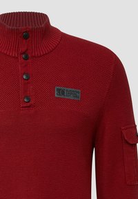 Czerwony sweter z dzianiny z prążkowanym kołnierzem, czterema czarnymi guzikami, naszywką na piersi z napisem "Elevated Casual Est. 1969" oraz kieszenią na rękawie.