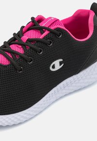 Zapatilla deportiva negra con parte superior de malla texturizada, acentos rosas, cordones negros y suela de goma blanca con un diseño estampado.