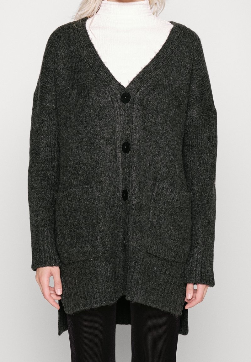 Femme portant un cardigan en tricot gris foncé boutonné avec deux poches avant sur un col roulé blanc et un pantalon noir.