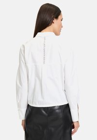 Blusa bianca in cotone a maniche lunghe con un elegante design sul retro, caratterizzata da una linea verticale di piccoli dettagli decorativi e polsini con bottoni.