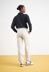 Fekete, hosszú ujjú, galléros ing, krémszínű, elasztikus mandzsettás melegítőnadrág, és fekete Nike sportcipő egy sárga padlón.